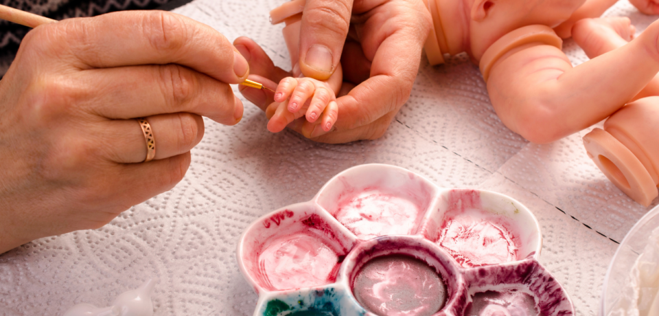 Vinyl reborn doll being hand painted with fine detail brush, showing realistic skin tone and craftsmanship
