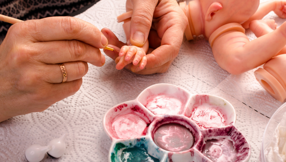 Vinyl reborn doll being hand painted with fine detail brush, showing realistic skin tone and craftsmanship